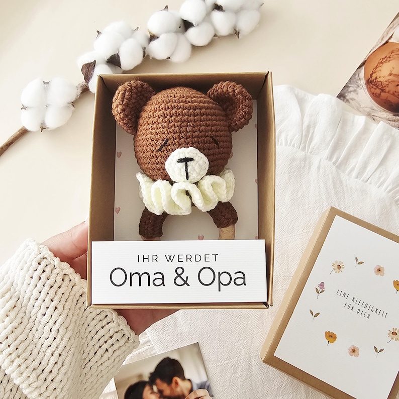 Puede incluir: Un sonajero de oso tejido a ganchillo en color marr&oacute;n con un cuello de volantes blanco dentro de una caja de regalo de cart&oacute;n. Una etiqueta blanca en la caja muestra el texto "IHR WERDET Oma & Opa" en negro.