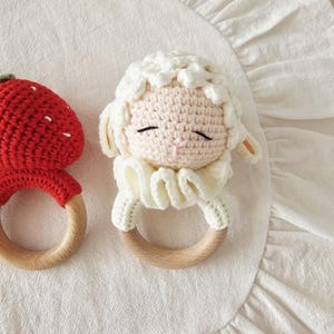 Puede incluir: Dos sonajeros de ganchillo con base de anillo de madera. Un sonajero tiene forma de fresa roja con semillas blancas bordadas. El otro sonajero tiene forma de oveja de color crema con una cabeza de lana texturizada y un dise&ntilde;o de cuello con volantes.