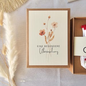 Puede incluir: Una caja de regalo rectangular de cart&oacute;n marr&oacute;n con una etiqueta de color crema que presenta una ilustraci&oacute;n en acuarela de flores silvestres en tonos naranja y marr&oacute;n. El texto EINE BESONDERE &Uuml;berraschung est&aacute; impreso en negro.