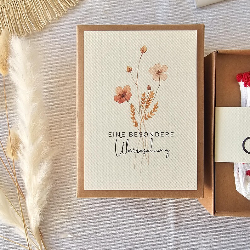 Puede incluir: Una caja de regalo rectangular de cart&oacute;n marr&oacute;n con una etiqueta de color crema que presenta una ilustraci&oacute;n en acuarela de flores silvestres en tonos naranja y marr&oacute;n. El texto EINE BESONDERE &Uuml;berraschung est&aacute; impreso en negro.