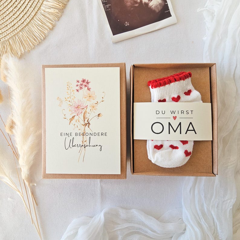Puede incluir: Una caja de regalo que contiene un par de calcetines blancos con peque&ntilde;os corazones rojos y borde rojo, envueltos en una banda de papel que dice DU WIRST OMA con un icono de coraz&oacute;n. Al lado, una tarjeta con una ilustraci&oacute;n floral y el texto EINE BESONDERE &Uuml;berraschung.