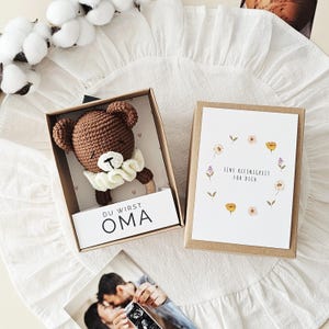 Puede incluir: Un sonajero de oso tejido a ganchillo marr&oacute;n con un cuello de volantes color crema dentro de una caja de cart&oacute;n. La caja presenta el texto DU WIRST OMA y una tarjeta separada con un dise&ntilde;o de corona floral y el texto EINE KLEINIGKEIT F&Uuml;R DICH.