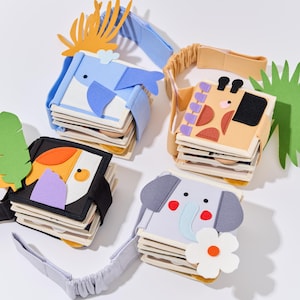 May include: Four colorful felt animal books with elastic straps. The books feature a whale, giraffe, toucan, and elephant. Each book has multiple pages and is decorated with felt details and a flower.