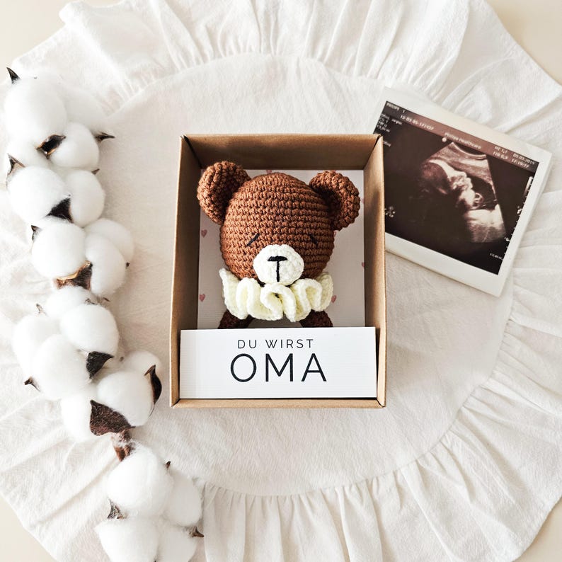 Puede incluir: Un oso de ganchillo marr&oacute;n con un cuello de volantes color crema se encuentra dentro de una caja de regalo de cart&oacute;n. Una tarjeta blanca en la caja presenta el texto DU WIRST OMA. El oso tiene ojos y nariz bordados, y la caja se presenta con algod&oacute;n.