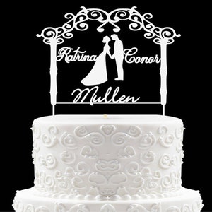 Può includere: Topper per torta bianco con una silhouette di una sposa e uno sposo in piedi sotto un arco. Il topper include i nomi "Katrina Connor Mullen".