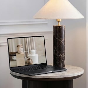 Może przedstawiać: Czarny laptop wyświetlający na ekranie obraz ceramicznych wazonów, ustawiony obok cylindrycznej lampy stołowej z ciemnobrązowego marmuru z białym, stożkowym abażurem z tkaniny. Na ekranie widoczny jest tekst LE BUREAU MOCKUP COLLECTION.