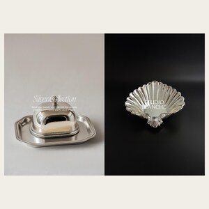 May include: Two silver serving pieces. A silver cloche with a domed lid sits on a silver tray. A silver shell-shaped dish with a fluted edge.