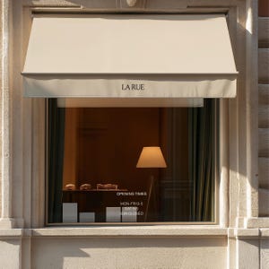 May include: A storefront window with a beige awning featuring the text LA RUE. The glass displays opening times: MON-FRI 9-5, SAT 9-5, SUN CLOSED. Inside, a floor lamp with a white shade stands next to a shelf with small items on a dark surface.