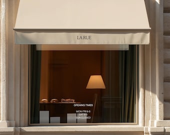 May include: A storefront window with a beige awning featuring the text LA RUE. The glass displays opening times: MON-FRI 9-5, SAT 9-5, SUN CLOSED. Inside, a floor lamp with a white shade stands next to a shelf with small items on a dark surface.
