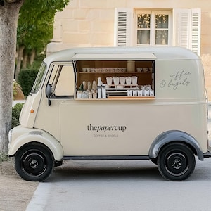 Puede incluir: Un camión de comida de café y bagels de color crema, con ruedas negras y una ventanilla lateral. El camión tiene el texto "thepapercup COFFEE & BAGELS" y "coffee & bagels" en escritura cursiva. La ventanilla muestra tazas de café y suministros.