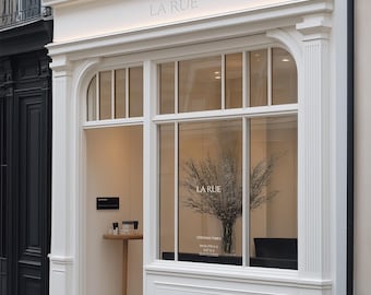 May include: A storefront with a white facade and large glass windows. The text LA RUE is displayed above the entrance and on the window. Inside, a tall vase holds dried branches. A small wooden table stands near the entrance with items on it.