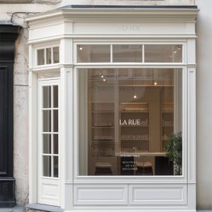 Può includere: Una vetrina color crema con un'ampia finestra in vetro e una porta a più riquadri. Sul vetro è riportata la scritta LA RUE e gli orari: MON-FRI 9-5, SAT 9-5, SUN CLOSED. L'interno mostra scaffali con prodotti e una piccola pianta.