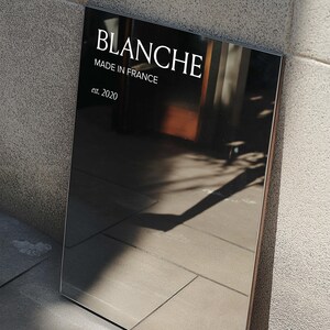 Op de afbeelding: Een rechthoekige zwarte spiegel met het woord "BLANCHE" in het wit, samen met "MADE IN FRANCE" en "est. 2020". De spiegel reflecteert de omgeving, waardoor een donker, glanzend oppervlak ontstaat.