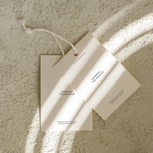 K&ouml;nnte beinhalten: Zwei cremefarbene Bekleidungsetiketten-Mockups mit dem Text "GARMENT TAG MOCKUP" und "PREMIUM MOCKUP WITH PSD SMART LAYERS". Die Etiketten sind mit einer wei&szlig;en Schnur verbunden und liegen auf einer strukturierten, hellen Oberfl&auml;che.