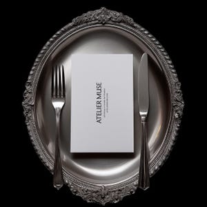 Op de afbeelding: Een zilverkleurige ovale schaal met sierlijke randen, een vork, een mes en een witte kaart met de tekst "ATELIER MUSE" in zwart. De kaart bevat ook de tekst "ETHICALLY MADE IN FRANCE" en een websiteadres. De achtergrond is zwart.