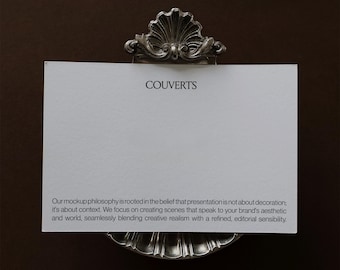 Könnte beinhalten: Eine rechteckige weiße Karte mit dem Wort COUVERTS in schwarzer Schrift oben und einem Absatz mit schwarzem Text unten, gehalten von einem silberfarbenen Metallständer mit einem dekorativen muschelartigen Design an den oberen und unteren Rändern.