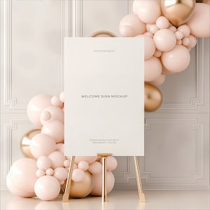 May include: A white welcome sign mockup on an easel with a gold base. The sign is surrounded by a cluster of pink and gold balloons.
