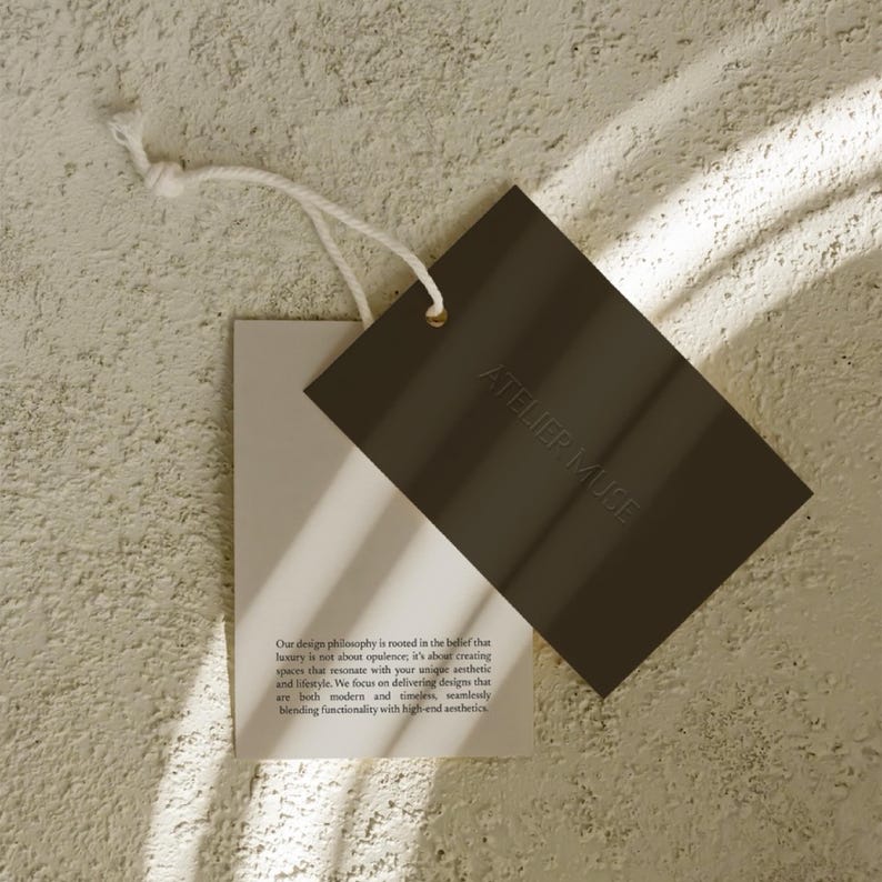 K&ouml;nnte beinhalten: Zwei rechteckige Anh&auml;nger, einer hell und einer dunkelbraun, h&auml;ngen an einer wei&szlig;en Schnur. Der dunkle Anh&auml;nger tr&auml;gt den gepr&auml;gten Text "ATELIER MUSE". Der hellere Anh&auml;nger enth&auml;lt Text, der die Designphilosophie der Marke beschreibt.