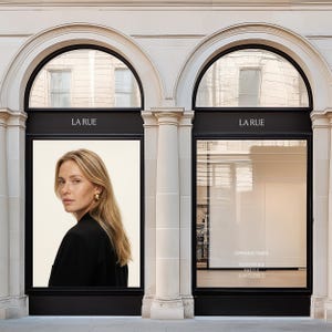 May include: A storefront with two arched windows framed in black, featuring the text LA RUE above each. The left window displays a portrait of a person with blonde hair wearing a black blazer and gold earrings. The right window shows opening times.