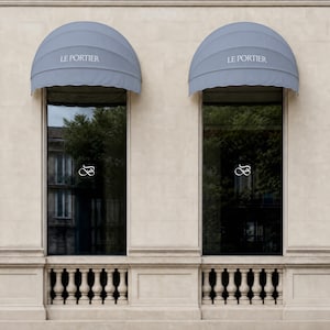 May include: Two tall rectangular windows set into a light beige stone wall, each topped with a rounded light blue fabric awning featuring the text LE PORTIER. The glass panes display a white infinity-style logo and reflect trees and buildings.