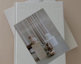 Puede incluir: Una pila de libros y un folleto sobre un fondo neutro. El libro superior es blanco con el título en relieve "THE KINFOLK HOME". El folleto presenta imágenes de cerámica y texto sobre diseño de interiores.