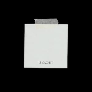 Puede incluir: Una nota cuadrada de papel blanco con el texto LE CACHET impreso en negro en la parte inferior. La nota está sujeta en la parte superior central con una tira de cinta adhesiva translúcida de color blanco roto sobre un fondo negro.