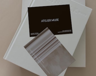 May include: A still-life image featuring two white hardcover books with embossed text, and a black business card with the text "ATELIER MUSE". A brochure with a beige and white design is also visible.