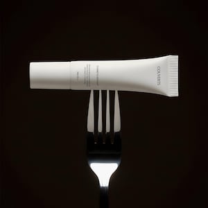 May include: A white cosmetic tube with a screw cap balanced horizontally on a silver fork. The tube features the text "COUVERTS" and "COSMETIC MOCKUP" along with "10ml / 0.34 fl. oz." printed in black against a solid, dark, and neutral background.