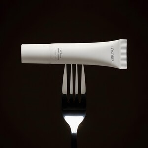May include: A white tube of "LONDRES" lip balm, resting on a silver fork against a black background. The tube has the words "Lip Balm, Baume à Lèvres" printed on it.