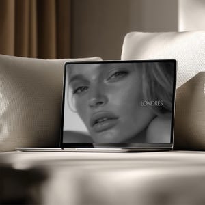 May include: A silver laptop displaying a black and white image of a woman's face with the word "LONDRES" in white text. The laptop is open and resting on a beige sofa, with a neutral-toned background.