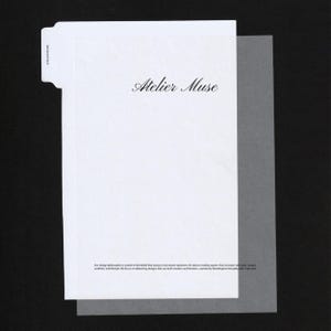 以下が含まれることがあります： エレガントな筆記体で「Atelier Muse」と書かれた白い紙の封筒。半透明のシートが封筒を部分的に覆っています。デザイン哲学が下部に印刷されています。背景は黒です。