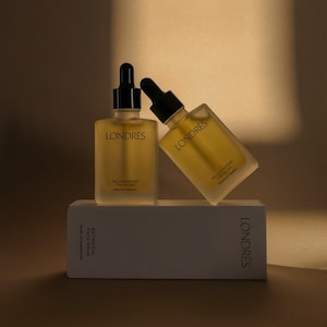 Puede incluir: Dos botellas de vidrio de aceite facial rejuvenecedor Londrés con cuentagotas negros, una vertical y otra inclinada. Las botellas contienen un líquido dorado y están sobre una caja blanca. El logotipo de Londrés es visible.