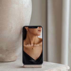 以下が含まれることがあります： アトリエミューズというジュエリーブランドのウェブサイトを表示している黒いスマートフォン。ウェブサイトには、金色のイヤリングと小さなペンダントのネックレスをつけたモデルが写っています。ウェブサイトのテキストには、「アトリエミューズ」と「スタッカブル。フランス製。ateliermuse.com」と表示されています。