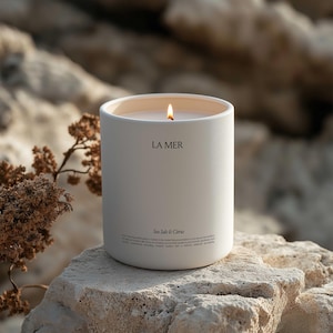 Könnte beinhalten: Eine brennende, cremefarbene Kerze mit dem Text "LA MER" und "Sea Salt & Citrus" auf der Vorderseite. Die Kerze steht auf einem hellen Felsen, mit einem verschwommenen natürlichen Hintergrund.