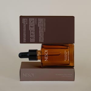May include: A brown glass bottle of NESOI Rejuvenating Facial Oil with a black dropper, on a brown box. The box below reads "Botanical Face Balm". Another brown box is above.