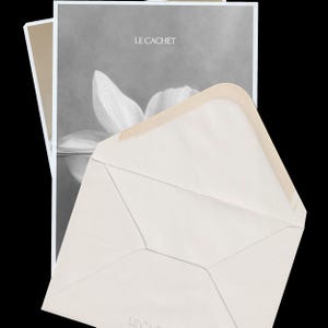 Puede incluir: Un sobre de color crema con solapa puntiaguda colocado frente a una tarjeta rectangular. La tarjeta presenta una imagen en escala de grises de una flor blanca con gotas de agua y el texto LE CACHET impreso en blanco en la parte superior.