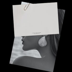 May include: A black and white image featuring a woman wearing a large, silver seashell-shaped earring. A white card with the text "ATELIER MUSE" and "ETHICALLY MADE IN FRANCE" is held above the woman's head, secured by a paper clip.