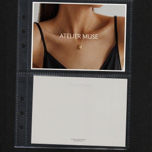 May include: A gold pendant necklace with a small, rounded rectangular charm. The necklace is displayed on a person wearing a black top. The text "ATELIER MUSE" is visible on the image.