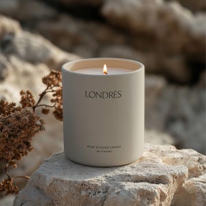 Puede incluir: Una vela blanca con una etiqueta marrón claro que dice "LONDRES ROSE SCENTED CANDLE Rose & Bergamot". La vela está encendida y se encuentra sobre una roca marrón claro.