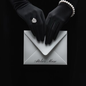 May include: A white envelope with the text "Atelier Muse" is held by gloved hands. A diamond ring and pearl bracelet are also visible. The background is black.