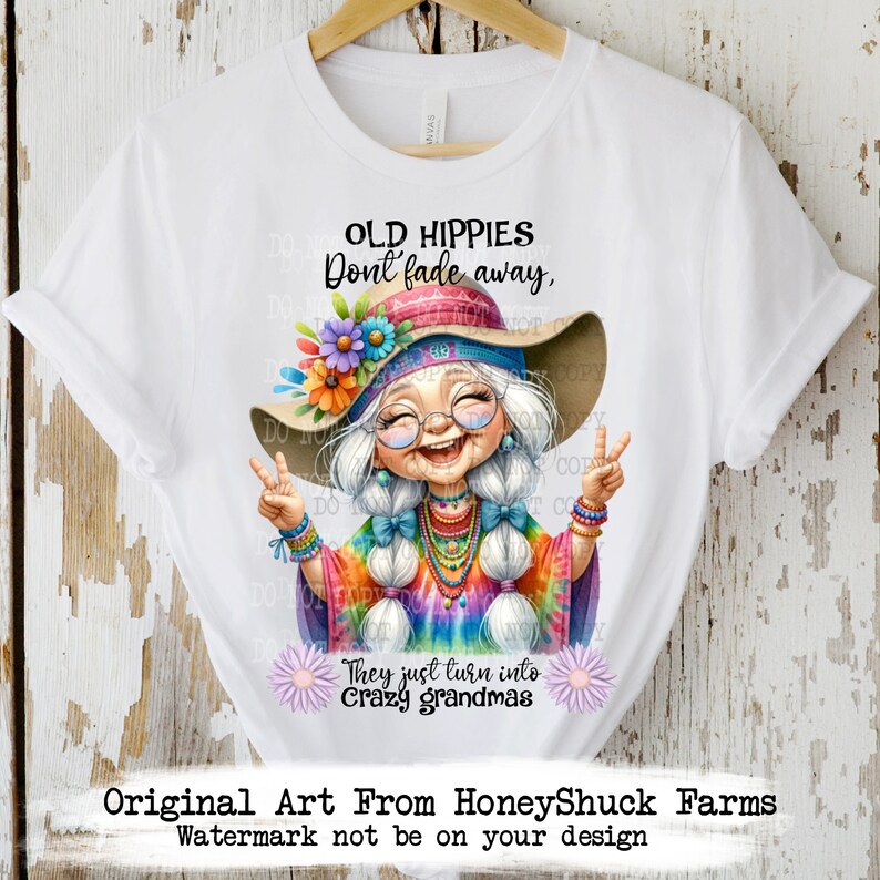 May include: A white t-shirt featuring a colourful illustration of a smiling woman with long white hair wearing a tie-dye shirt, a flower crown, and a peace sign necklace. The text "Old Hippies Don't Fade Away, They Just Turn Into Crazy Grandmas" is printed on the shirt.