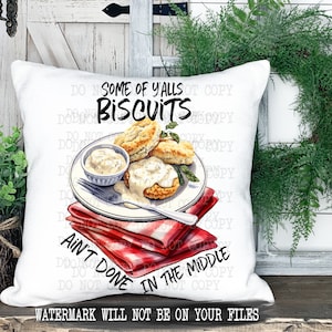 Op de afbeelding: Een wit kussen met een rood-wit geruit servet en een bord met koekjes en jus. De tekst op het kussen luidt "Some of y'alls biscuits ain't done in the middle."