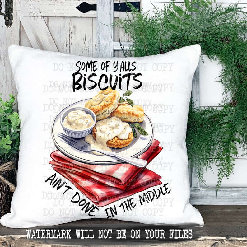 Op de afbeelding: Een wit kussen met een rood-wit geruit servet en een bord met koekjes en jus. De tekst op het kussen luidt "Some of y'alls biscuits ain't done in the middle."