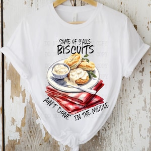 Op de afbeelding: Wit T-shirt met een afbeelding van biscuits en jus op een rood-wit geblokte servet. De tekst "Some of y'alls biscuits ain't done in the middle" staat op het T-shirt gedrukt.