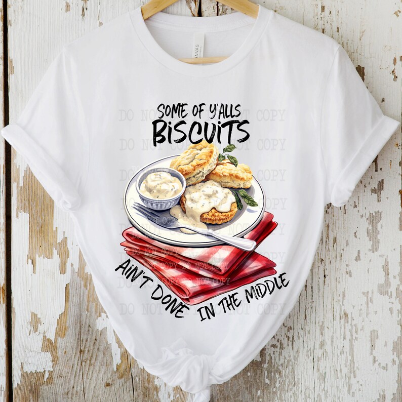 Op de afbeelding: Wit T-shirt met een afbeelding van biscuits en jus op een rood-wit geblokte servet. De tekst "Some of y'alls biscuits ain't done in the middle" staat op het T-shirt gedrukt.