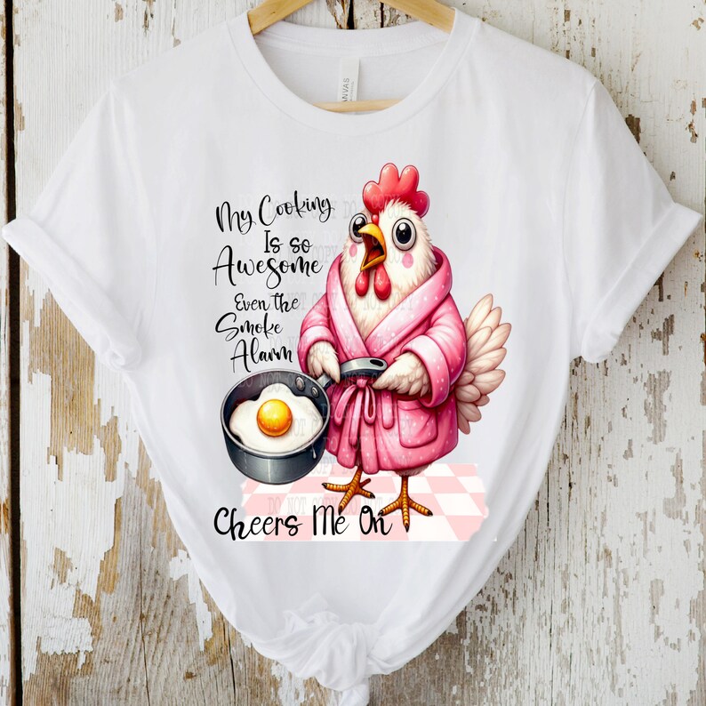 Pu&ograve; includere: Una maglietta bianca con un pollo rosa che indossa una vestaglia rosa e tiene una padella con un uovo fritto. Il testo sulla maglietta recita "My Cooking Is So Awesome Even The Smoke Alarm Cheers Me On".