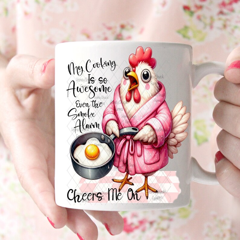 Pu&ograve; includere: Una tazza in ceramica bianca con un'illustrazione rosa e bianca di una gallina che indossa una vestaglia rosa e tiene una padella con un uovo fritto. Il testo sulla tazza recita "My Cooking Is So Awesome Even The Smoke Alarm Cheers Me On".