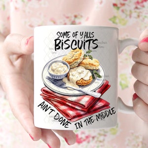 Op de afbeelding: Een witte keramische mok met een rood-wit geruite servet, een bord met biscuits en jus, en de tekst "Some of y'alls biscuits ain't done in the middle".