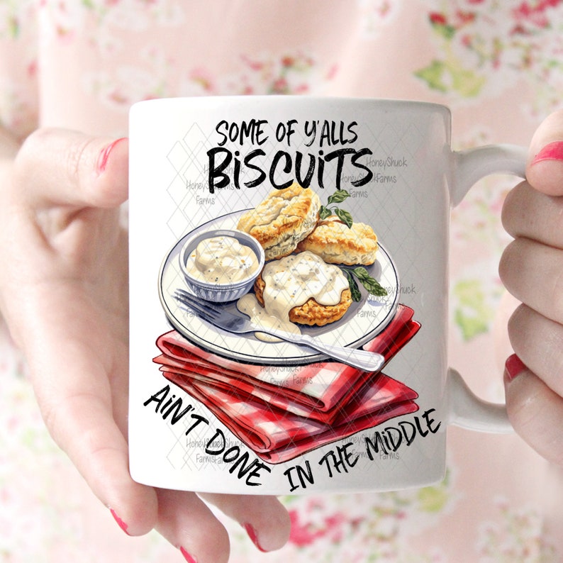 Op de afbeelding: Een witte keramische mok met een rood-wit geruite servet, een bord met biscuits en jus, en de tekst "Some of y'alls biscuits ain't done in the middle".