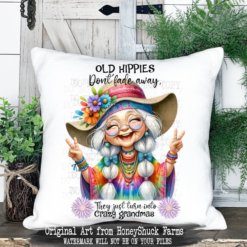 May include: A white cushion featuring a colourful illustration of a smiling woman with long white hair wearing a tie-dye shirt, a flower crown, and a wide-brimmed hat. The text "Old Hippies Don't Fade Away, They Just Turn Into Crazy Grandmas" is printed on the cushion.